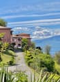 The image shows a group of multi-story residential buildings with a Mediterranean-style architecture, surrounded by lush greenery and overlooking a scenic lake or body of water. The buildings have terracotta-colored roofs and are set against a backdrop of