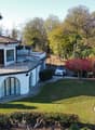 Two-story white villa with tiled roof, large balcony, garage, swimming pool, surrounded by lush greenery and trees