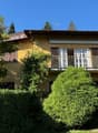 2-story yellow house with a balcony, surrounded by lush greenery and trees