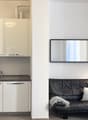 The image shows a small studio apartment with a kitchenette and a living area. The kitchenette has white cabinets, a sink, and a dishwasher. The living area has a black leather sofa, a large wall mirror, and a floor lamp.