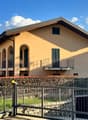 2-story yellow stucco house with arched windows, balconies, and a metal roof. The house is surrounded by a metal fence and landscaping with flowers and shrubs.