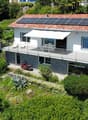 2-story modern house with a tiled roof, solar panels installed on the roof, a balcony or terrace, and surrounded by lush greenery and palm trees.