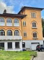 3-story yellow house with arched windows, decorative trim, and a balcony. The house has a garage and is surrounded by trees and greenery.