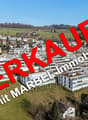 The image shows an aerial view of a town with a mix of residential and commercial buildings. The town is situated in a hilly, forested area with a clear blue sky in the background. The buildings appear to be a mix of modern and traditional architectural s