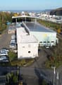 An industrial facility with a large parking area with multiple vehicles parked. A warehouse with a solar panel roof is situated on the property. Several buildings and trees are in the distance.