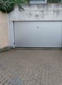 A concrete garage with two doors, one large and one small, and a small paved area in front.