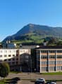 2 building structures, multiple cars parked, green grass and trees, view of a mountain