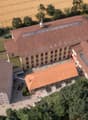 This image shows an aerial view of a large, multi-story brick building with a red tile roof. The building appears to be surrounded by trees and greenery, with a paved courtyard or plaza in front of it. There are several vehicles parked in the area around