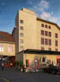 3-story building with arched windows, red trims, entrance with large canopy, sign reading FLOOS MAGAZINE, multiple parked cars
