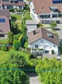 House with green roof, car parked in front, garden, solar panels, large grassy lawn, residential area, other houses and cars visible in the background