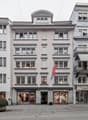 A multi-story building with a classical architectural style, featuring arched windows, decorative moldings, and a central pediment. The building has several retail stores on the ground floor, including 'Des petits hauts' and 'Blue Lemon'. A Serbian flag i