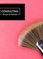 The image shows a black and white logo for 'GEWERBE CONSULTING Kruse & Partner' against a bright pink background. There is also a large makeup brush visible in the foreground.