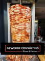 A large piece of meat, possibly chicken, is being cooked on a vertical rotisserie. The meat is sliced into thin layers and is being cooked with a red sauce. The meat is being cooked in a commercial kitchen with a stainless steel oven and a refrigerator in