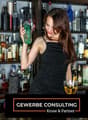 A woman in a black dress holding a green cocktail shaker in front of a bar shelf with various liquor bottles