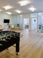 Large open office space with hardwood floors, white walls, and large windows. The space includes a foosball table, a dining table with chairs, and various potted plants. There is also a kitchen area visible through the doorway.