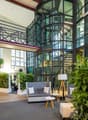 modern lobby, gray couch, white tables, large floor lamps, white wall, gray flooring, large plants, glass windows, staircase