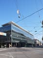 Large glass and steel commercial building with Credit Suisse branding, surrounded by historic ornate buildings, tram lines, and pedestrians on the street