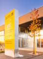 Modern commercial building, yellow signboard, glass facade, entrance with automatic sliding doors, glass wall, tree in front, brick walkway, paved entrance area