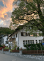 white facade house with brown wooden shutters, balcony on first floor, red roof, mountain in background