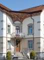 white house, brown roof, multiple windows, large entrance with steps, balcony