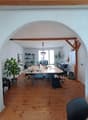 large room with wooden floor, two long tables, office chairs, desk, plant, desk lamp, framed picture, black bag, trolley, ceiling lamp