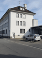 White building, two stories, multiple windows, parking in front, cars parked under carport.