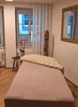 Room contains a chair, a table with a flower vase, a small bed with a fur blanket, and a large mirror
