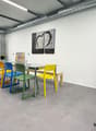 Open room with dining table and chairs. Light gray floors, white walls. Kitchen area with microwave, cabinets, bottles, kettle and a coffee machine. Bar stools next to a table with books and a framed art.