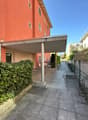Main entrance covered by a canopy, orange colored building, large greenery bush, pathway leading to the entrance, chain link fence on the side