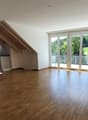 Spacious open-plan living area with hardwood flooring, large windows providing ample natural light, and access to a balcony or terrace overlooking a green outdoor area. The kitchen is integrated into the living space and features modern appliances.