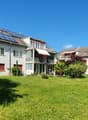 2 story apartment house with solar panels on the roof, surrounded by grass and plants, balconies, awnings, red shutters