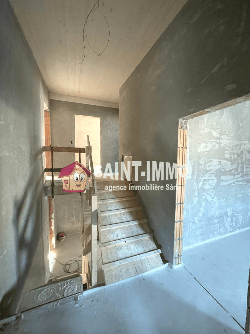 The image shows the interior of a building under construction or renovation. The walls are unfinished, with drywall or plaster visible. There is a wooden staircase leading to an upper level, and a small decorative house-shaped object can be seen on the st
