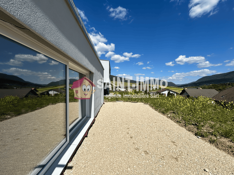 2-story modern house with large windows, gray exterior, gravel driveway, surrounded by green landscaping and mountains in the background