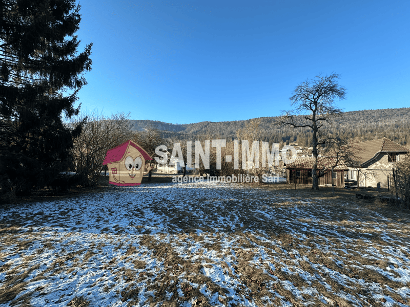 A plot of land covered in snow, with trees, a house in the distance, and mountains in the background.