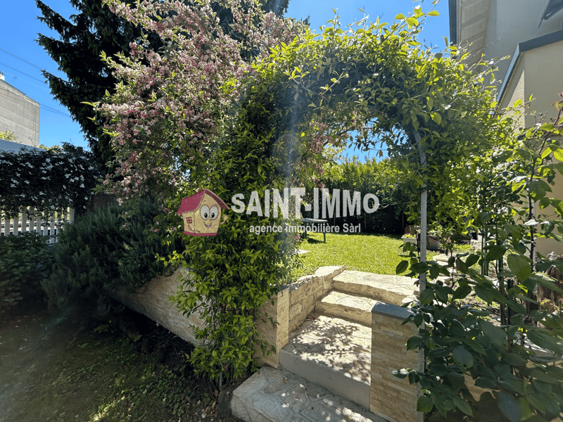 The image shows the entrance to a real estate office called 'Saint-Immo Agence Immobiliere Sarl'. The entrance is surrounded by lush greenery, including flowering trees and shrubs. There is a small stone pathway leading up to the entrance, and the office