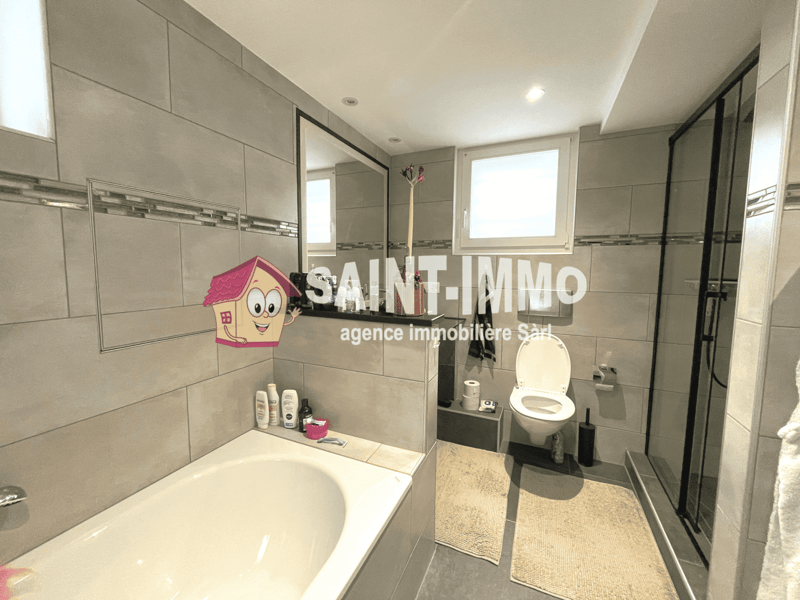 Spacious bathroom with grey tiled walls, a bathtub, toilet, and vanity with storage. There is a washing machine visible in the room.