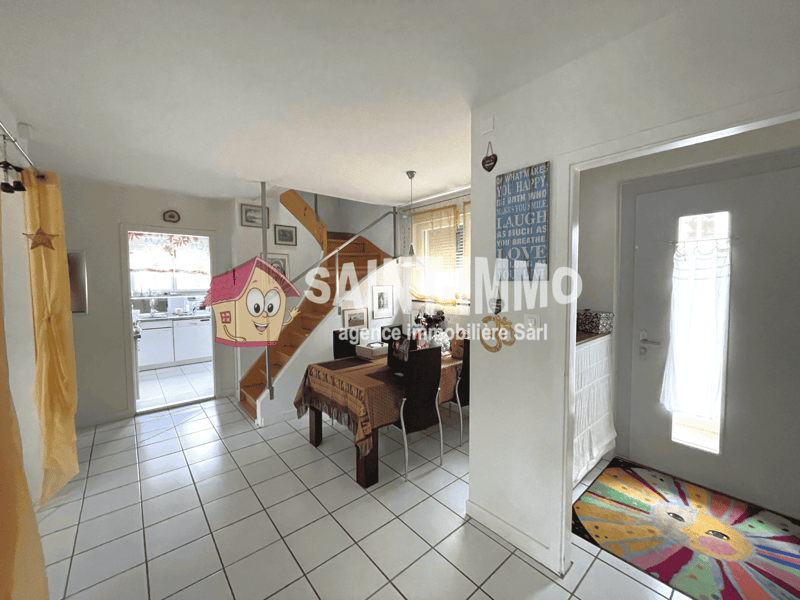 Open-concept kitchen and dining area with tiled flooring, white walls, wooden furniture, and decorative wall art. The kitchen area is visible through the doorway, and a dishwasher can be seen in the kitchen.