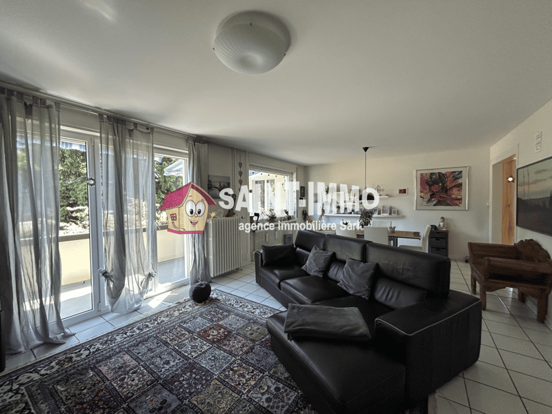 Spacious living room with large windows, black leather sofa, patterned rug, and a ceiling light fixture.