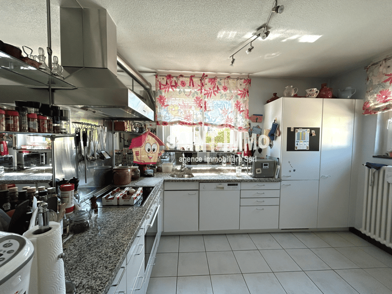 Spacious kitchen with granite countertops, stainless steel appliances, cabinets, and a decorative house-shaped item on the counter. The kitchen has a bright and colorful floral curtain and appears well-equipped.