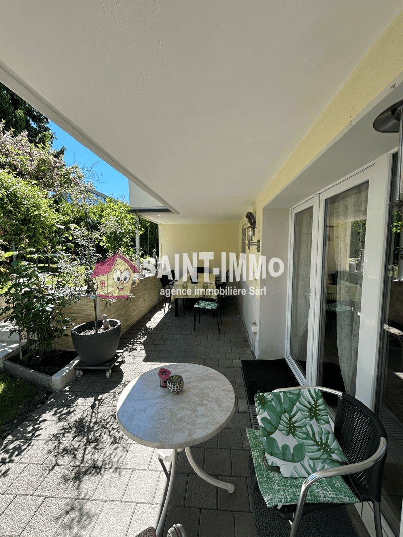 Covered patio with tiled flooring, a round table, chairs, and potted plants. The patio overlooks a lush, green garden area.