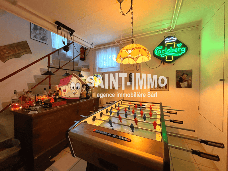 The image shows a game room with a foosball table, a Carlsberg neon sign, a stained glass lamp, and various decorative items and memorabilia on the shelves. The room has a cozy and lively atmosphere.
