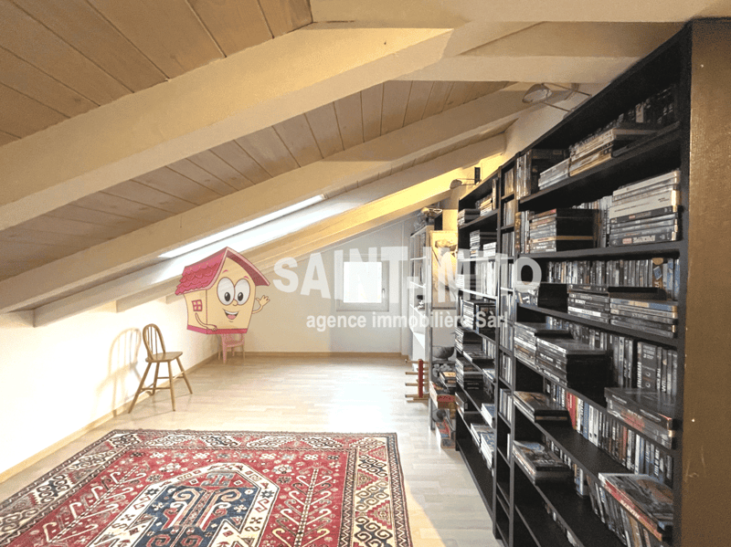 Attic space with wooden beams, shelves filled with books and media, a rug on the floor, and a cartoon house logo on the wall indicating this is the office of a real estate agency called Saint Remo.