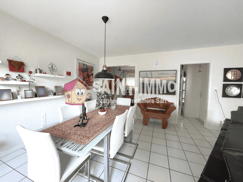 Open-concept living room and dining area, white walls, tile flooring, dining table with chairs, sofa, decorative shelves with artwork and accessories, pendant light fixture