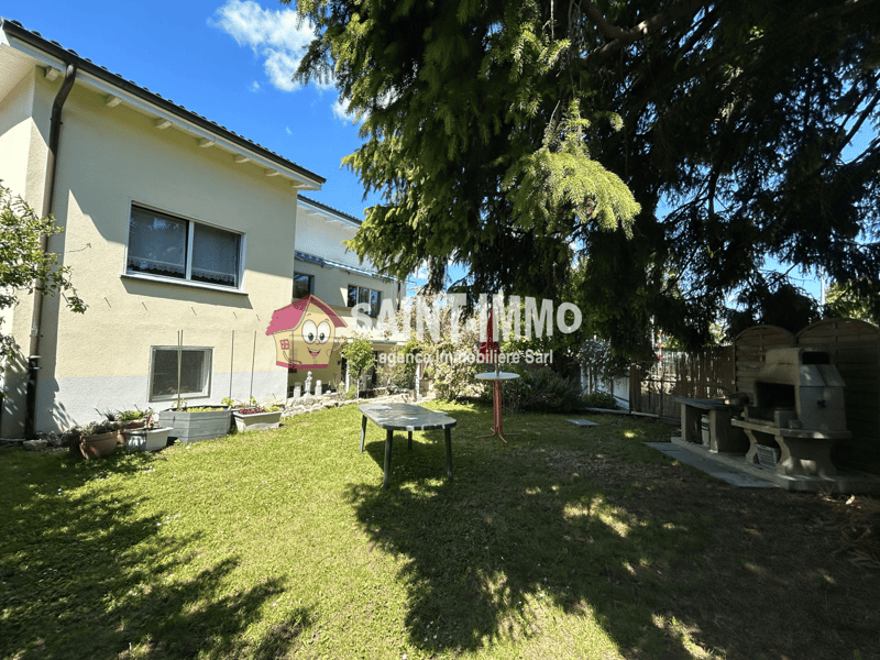2-story house with a yellow exterior, large windows, and a well-maintained garden with a grassy lawn, trees, and outdoor furniture. The property appears to have a parking area or driveway.