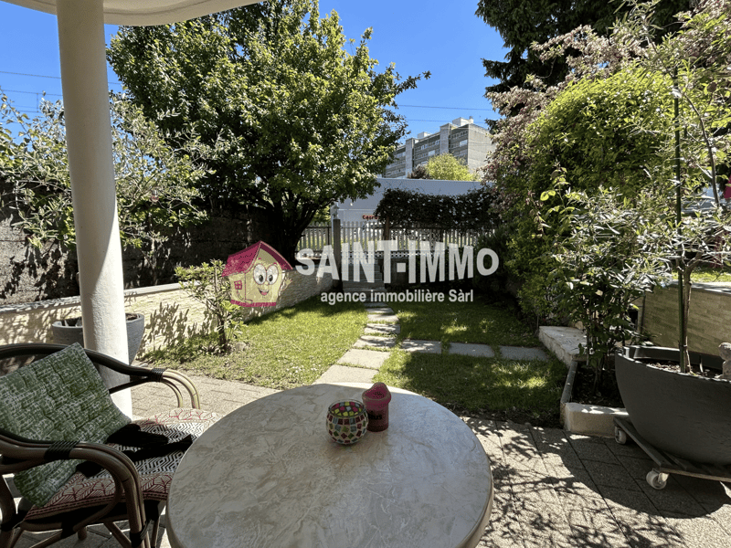 Outdoor patio area with a table and chairs, surrounded by lush greenery and trees. In the background, there is a sign for the real estate agency 'Saint-Immo' and a residential building.