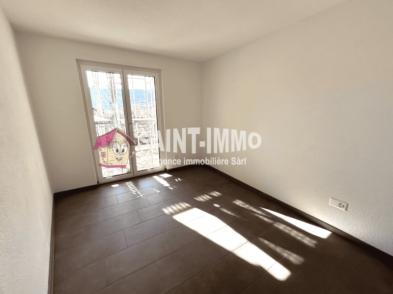 Empty room with hardwood flooring, white walls, and large windows providing natural light. The room appears to be a living or bedroom space.