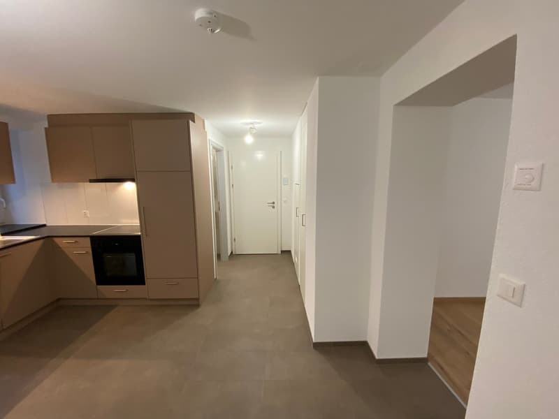 Empty hallway with white walls, gray floor, brown cupboards, open doorway, light switch, open doorway with lamp