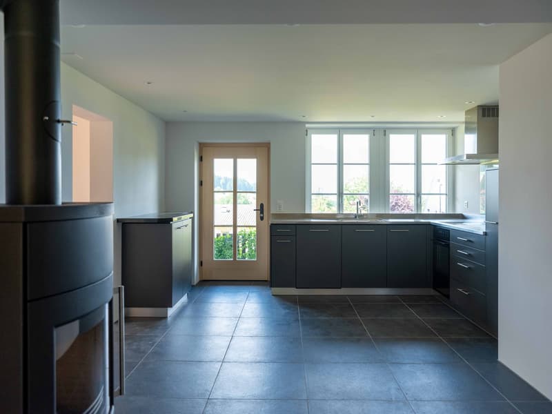 white walls, large windows, wood door, black tiled floor, gray cabinets, stainless steel countertops, wooden fireplace, stainless steel exhaust hood