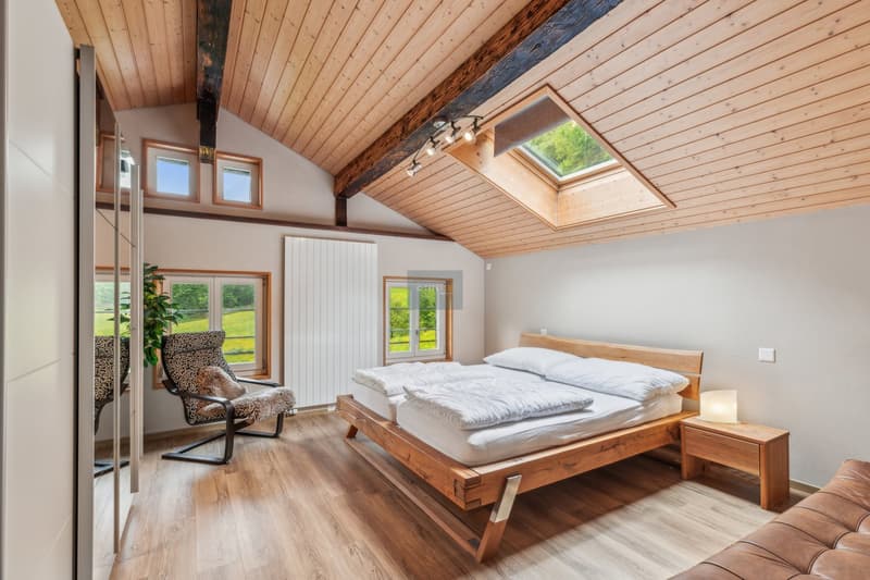 Spacious bedroom with vaulted wooden ceiling, large windows, wooden bed frame, armchair, and hardwood flooring