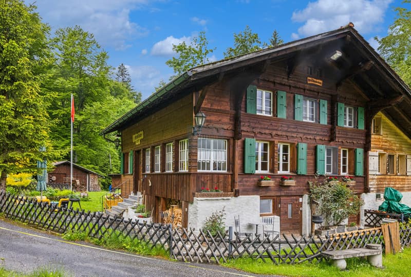 Two-story wooden chalet-style house with green shutters, surrounded by lush greenery and a wooden fence. The house has a covered porch and appears to be in a rural, forested setting.